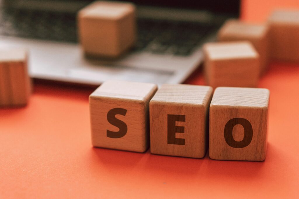 THREE WOODEN CUBES ARE PLACED ON A TABLE WHICH THE CUBES CONTAINS LETTERS "SEO".SOME OTHER WOODEN CUBES ARE IN THE BACKGROUND AND FEW OF THEM PLACED IN A LAPTOP.THE TABLE COLOUR IS ORANGE.