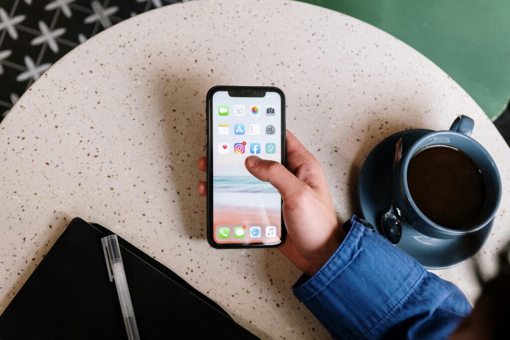 A CUP OF BLACK TEA,A WHITE PEN PLACED ON A BLACK POUCH PLACED ON A ROUND TABLE A GUYS WHITE RIGHT HAND WEARING BLUE SHIRT IS USING HIS MOBILE.